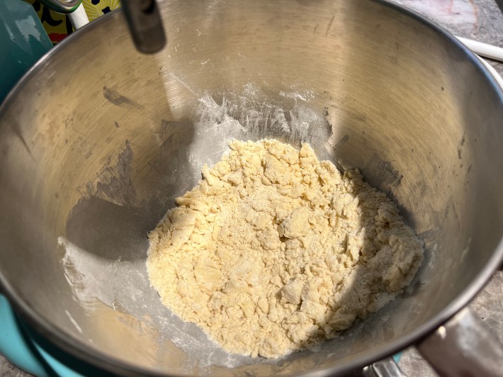 A metal mixing bowl holds a crumbly mixture of flour, sugar, egg, custard powder, and vanilla extract.