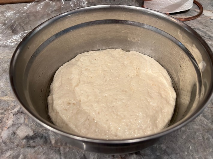 A metal mixing bowl holds a large pale blob of risen bread dough.