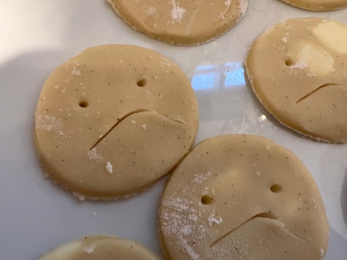 Close-up of cut ovals of raw cookie dough, with frowning faces cut into them with a chopstick and a paring knife.