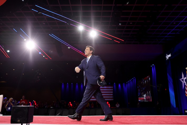 Ron DeSantis crosses a stage at CPAC, wearing heavy black boots. His weight is well forward of his back, right foot, but his right heel is on the ground, as if he is doing a calf stretch. The angle of his lower leg looks off.