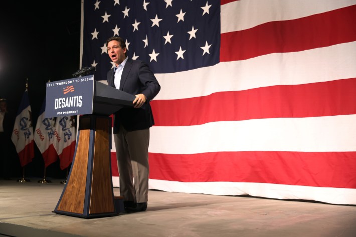 Ron DeSantis stands at a podium during a campaign rally. The heel of his left shoe is oddly angled.