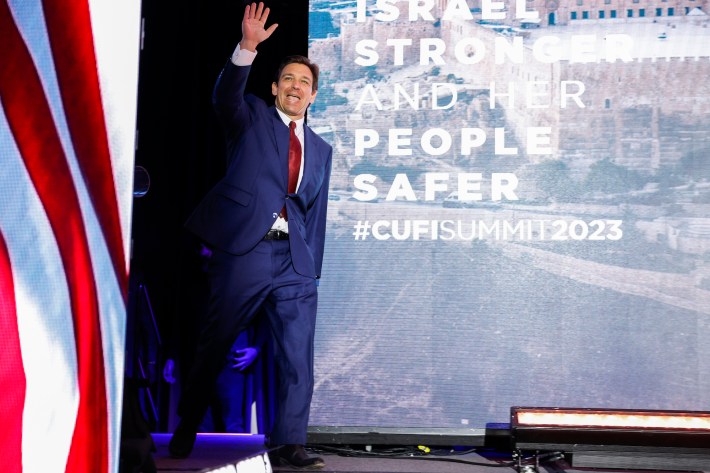 Ron DeSantis waves as he takes the stage at a summit in Virginia. His left leg appears to have an inexplicable bend roughly 12 inches above the floor, well below where his knee should be.