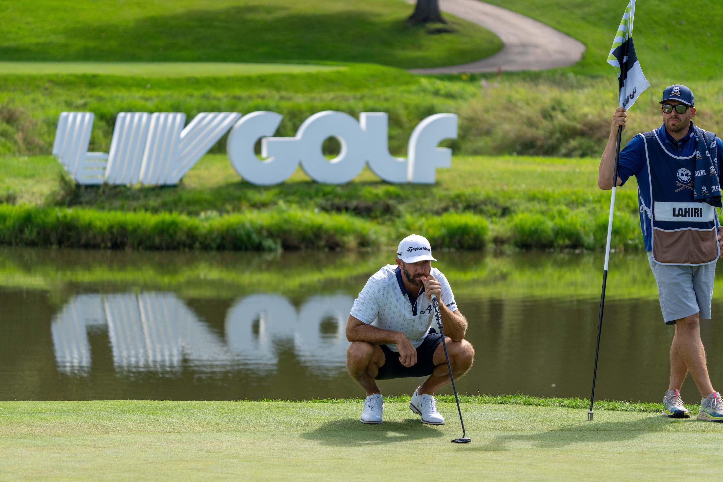 Dustin Johnson crouches on the green during a LIV Golf event, with a large "LIV GOLF" sign in the background. He is wearing shorts.