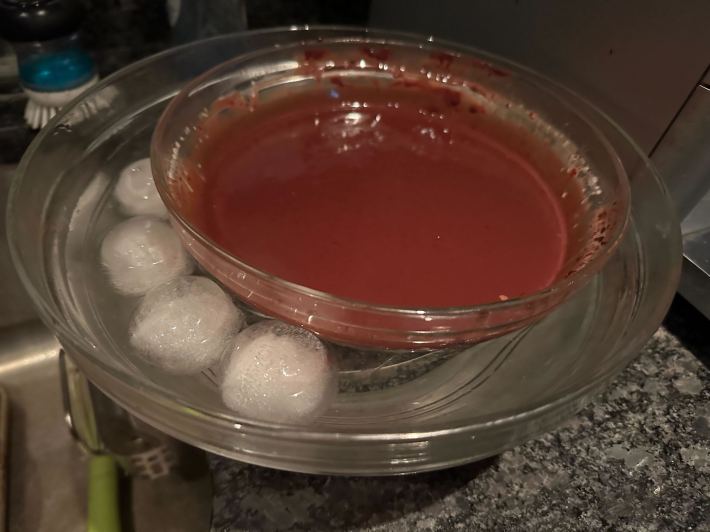 A glass bowl filled nearly to the brim with warm ganache, inside a larger glass bowl filled with ice water.