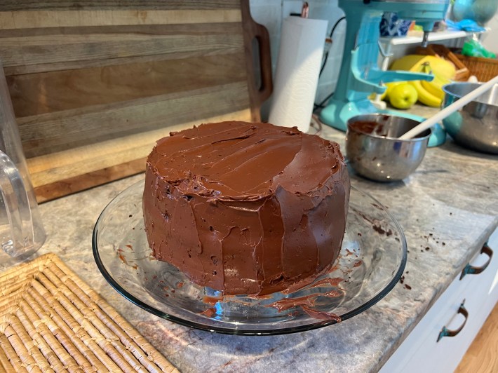 A reasonably well-iced two-tier cake sits on a glass cake stand, very nearly finished.