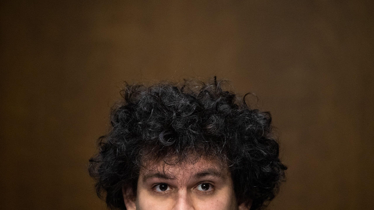 Samuel Bankman-Fried, founder and CEO of FTX, testifies during a Senate Committee on Agriculture, Nutrition and Forestry hearing about "Examining Digital Assets: Risks, Regulation, and Innovation," on Capitol Hill in Washington, DC, on February 9, 2022. The photo is stylized. His big poofy hair is mostly what the photo is of, plus the top half of his face. The canvas behind him is ugly and Brown, kind of in a gradient.