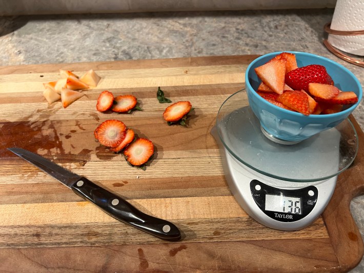 A small bowl of sliced strawberries measures 136 grams on a food scale, next to a pile of discarded strawberry stems and cores.