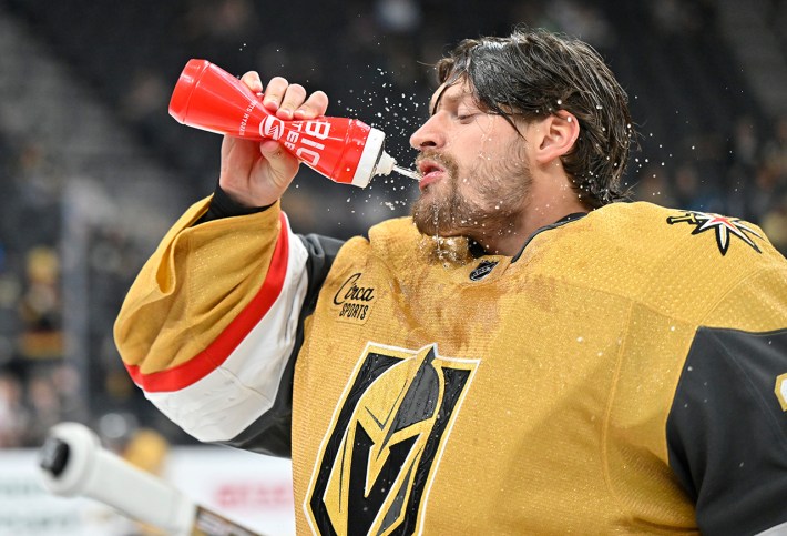 LAS VEGAS, NEVADA - OCTOBER 03: Adin Hill #33 of the Vegas Golden Knights warms up prior to a game against the San Jose Sharks at T-Mobile Arena on October 03, 2023 in Las Vegas, Nevada. He's squirting a red water bottle into his face and wearing the puke-yellow jersey of the Knights.