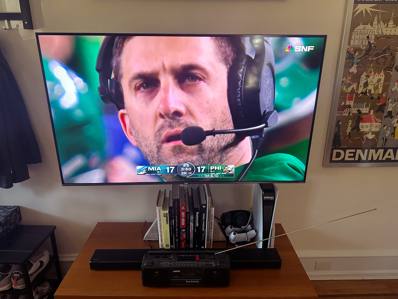 A photo of a television with a Dolphins/Eagles game on it, with Nick Sirianni's face filling the screen. Below it is a media cabinet, and there are some books and a PS5 behind a sound bar. In front of that sound bar is a small 1980s boombox.