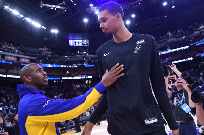 Victor Wembanyama of the San Antonio Spurs absolutely towers over Golden State's Chris Paul as they greet each other prior to a preseason game.
