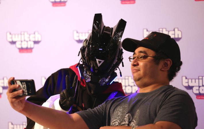 streamer girl_dm_ , wearing a black outfit with a helmet, poses with a fan for a selfie at TwitchCon 2023 in Las Vegas