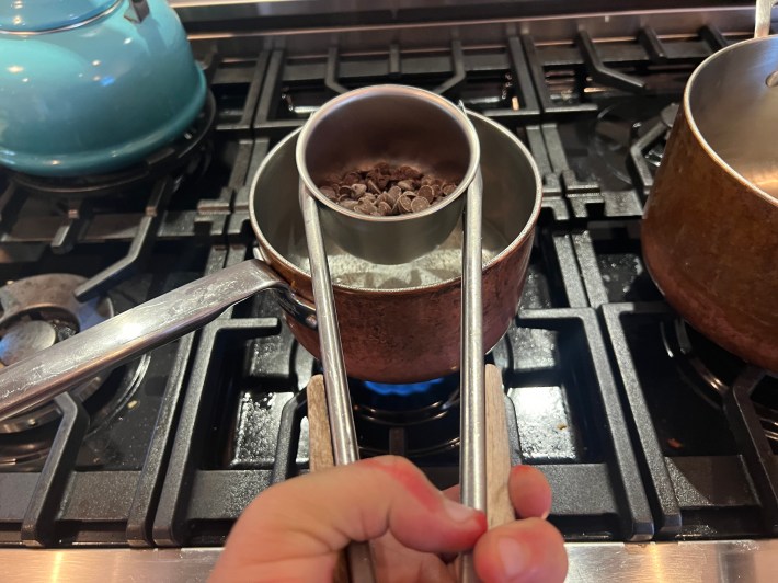 Semi-sweet chocolate chips in a small metal mixing bowl, held via tongs over a saucepan of simmering water.