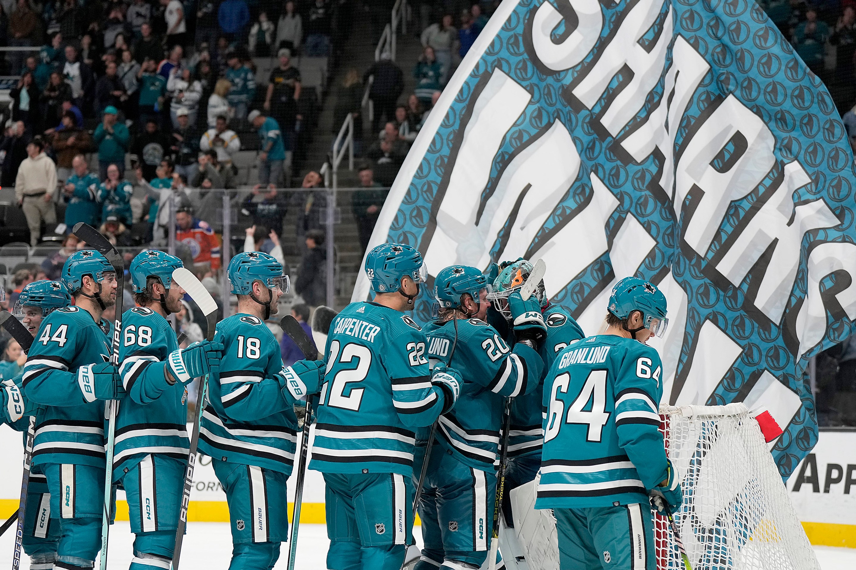 The San Jose Sharks celebrate with goalie Mackenzie Blackwood in front of a giant banner reading SHARKS WIN.