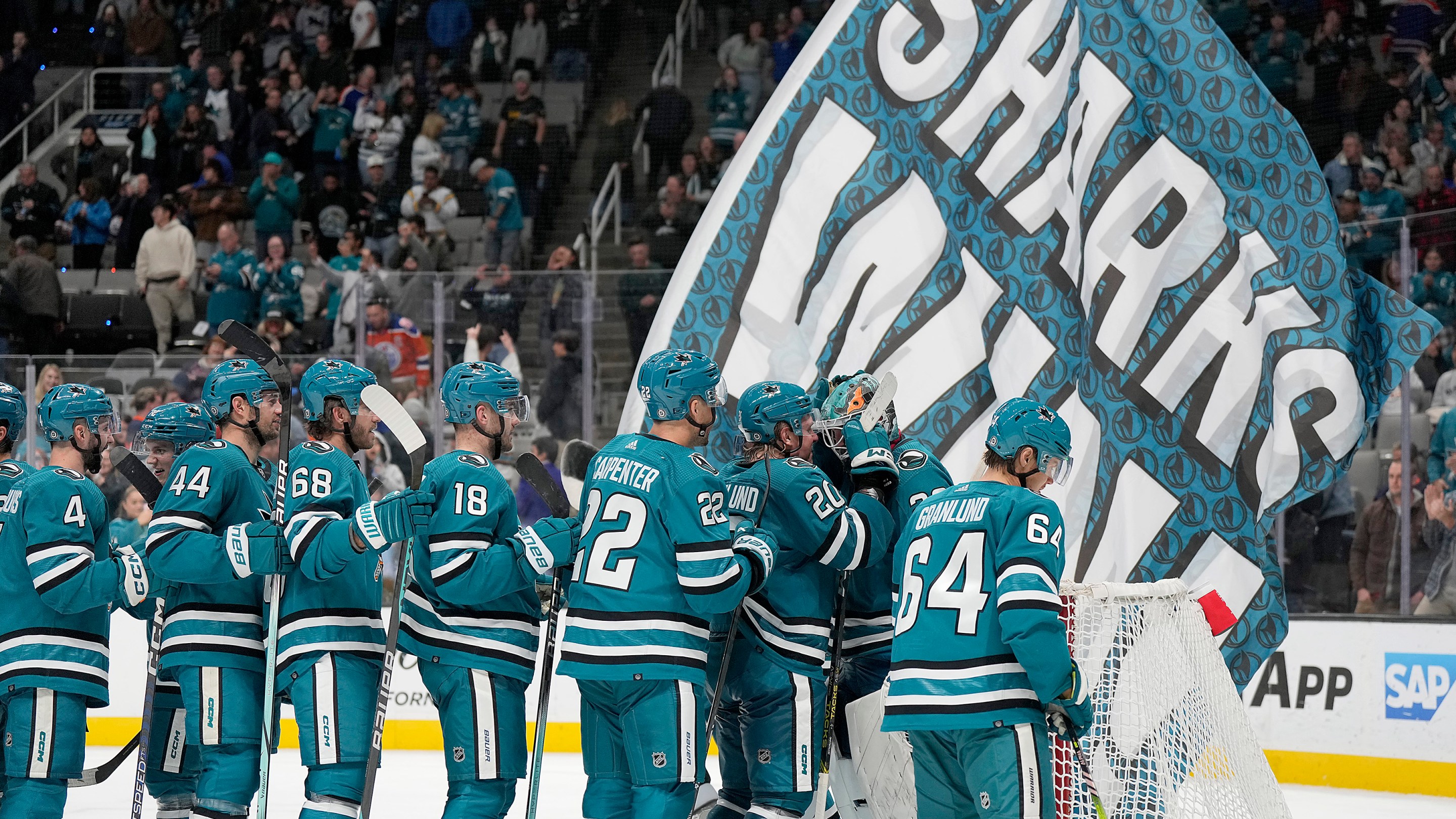 The San Jose Sharks celebrate with goalie Mackenzie Blackwood in front of a giant banner reading SHARKS WIN.
