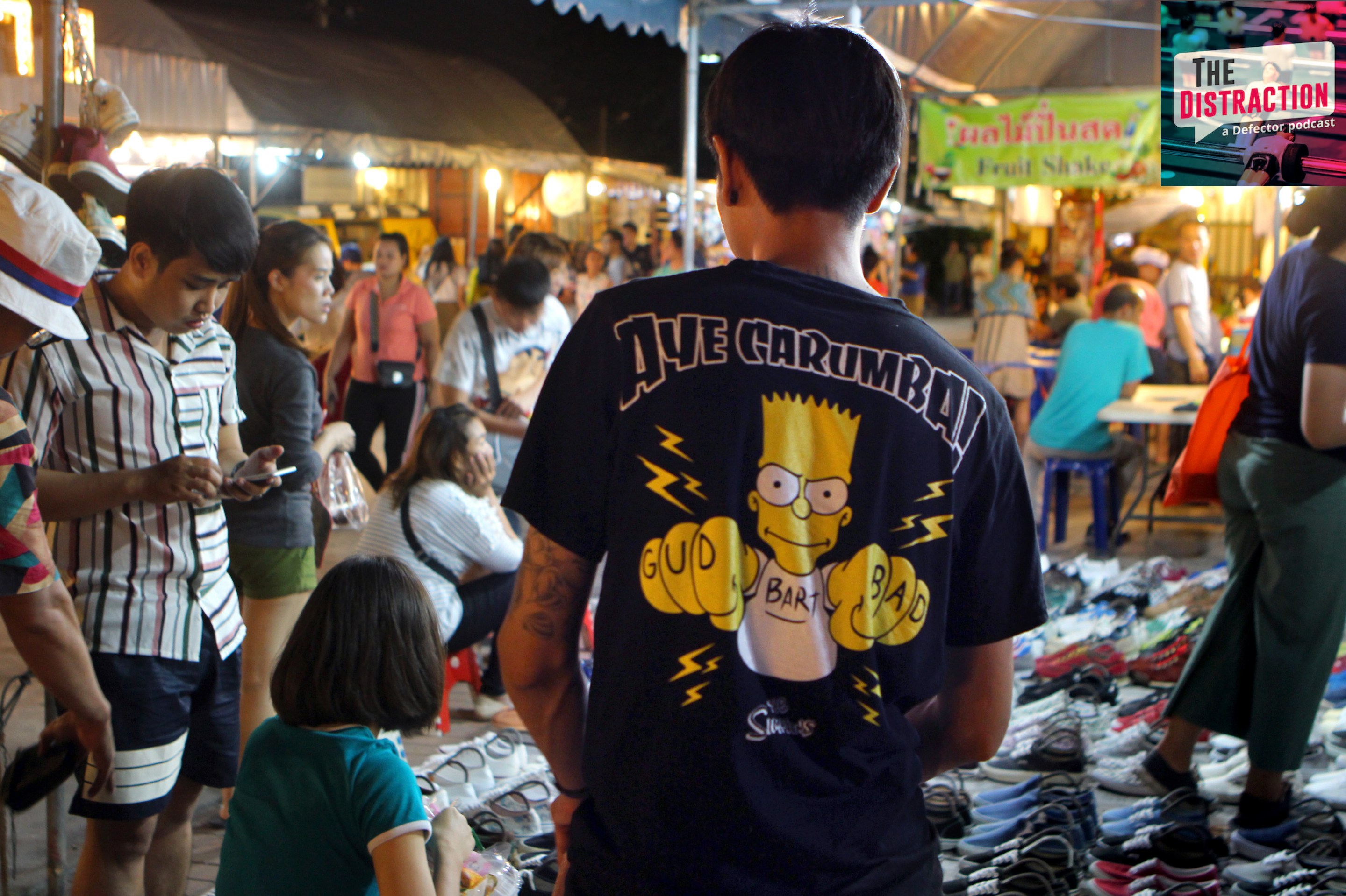 A man wears a bootleg Bart Simpson t-shirt—black, with the words Ay Carumba (sic) in heavy metal font over an image of Bart displaying GUD and BAD finger tattoos—on the streets of Bangkok, in 2018.