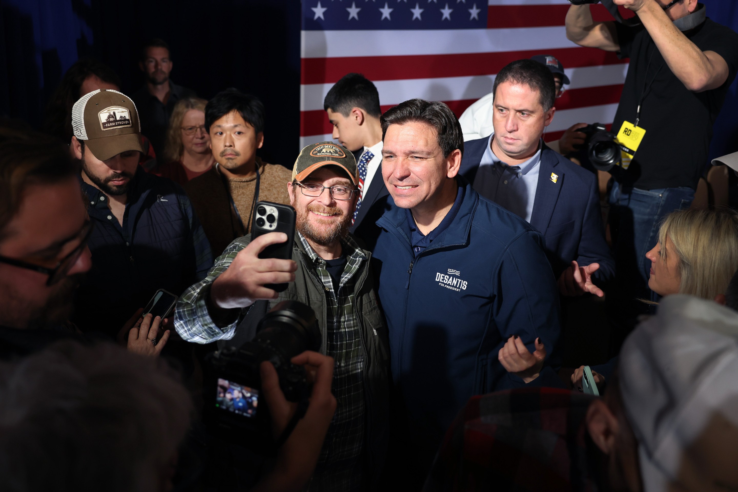 Ron DeSantis poses with a supporter, or anyway with a guy in a baseball hat, in Newton, Iowa in December of 2023. He is smiling in a very unsettling way.