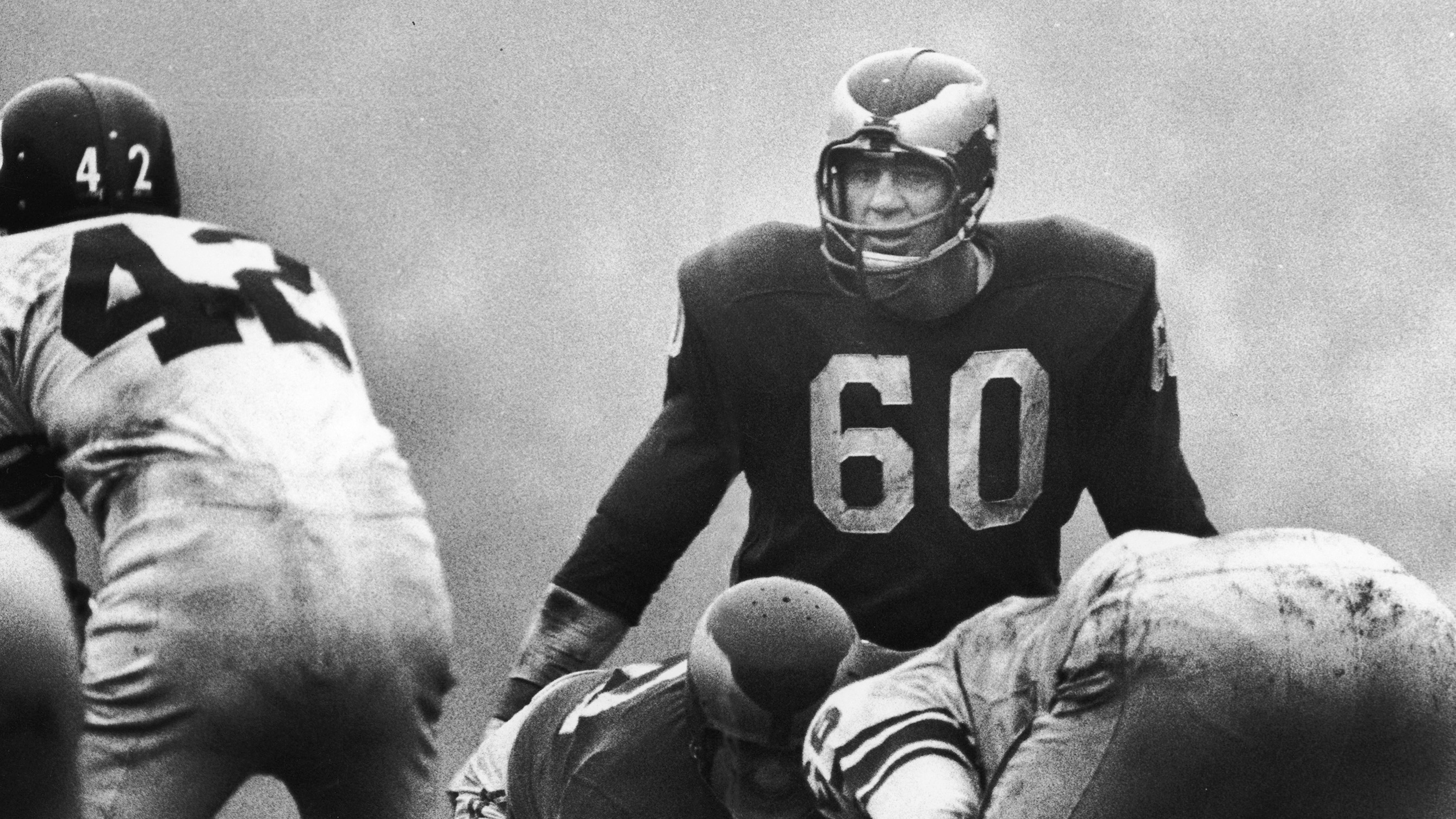 American football player Chuck Bednarik (#60) of the Philadelphia Eagles eyes his opponants as Charlie Conerly (#42), quarterback for the New York Giants prepares to take the ball during a game, late 1950s or early 1960s.