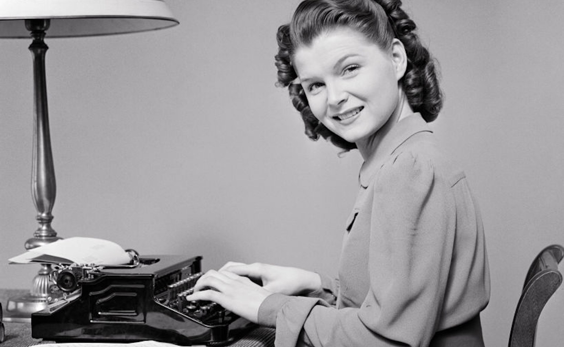 1940s Woman Seated At Home Desk Using Portable Typewriter Head Turned Looking At Camera Smiling.