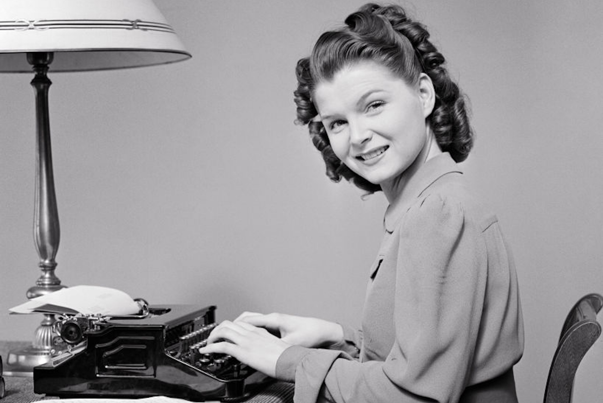 1940s Woman Seated At Home Desk Using Portable Typewriter Head Turned Looking At Camera Smiling.