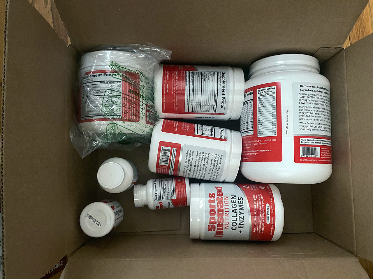 A box of Sports Illustrated supplements. White bottles of various sizes. The most visible bottle is Sports Illustrated Nutrition: Collagen + Enzymes