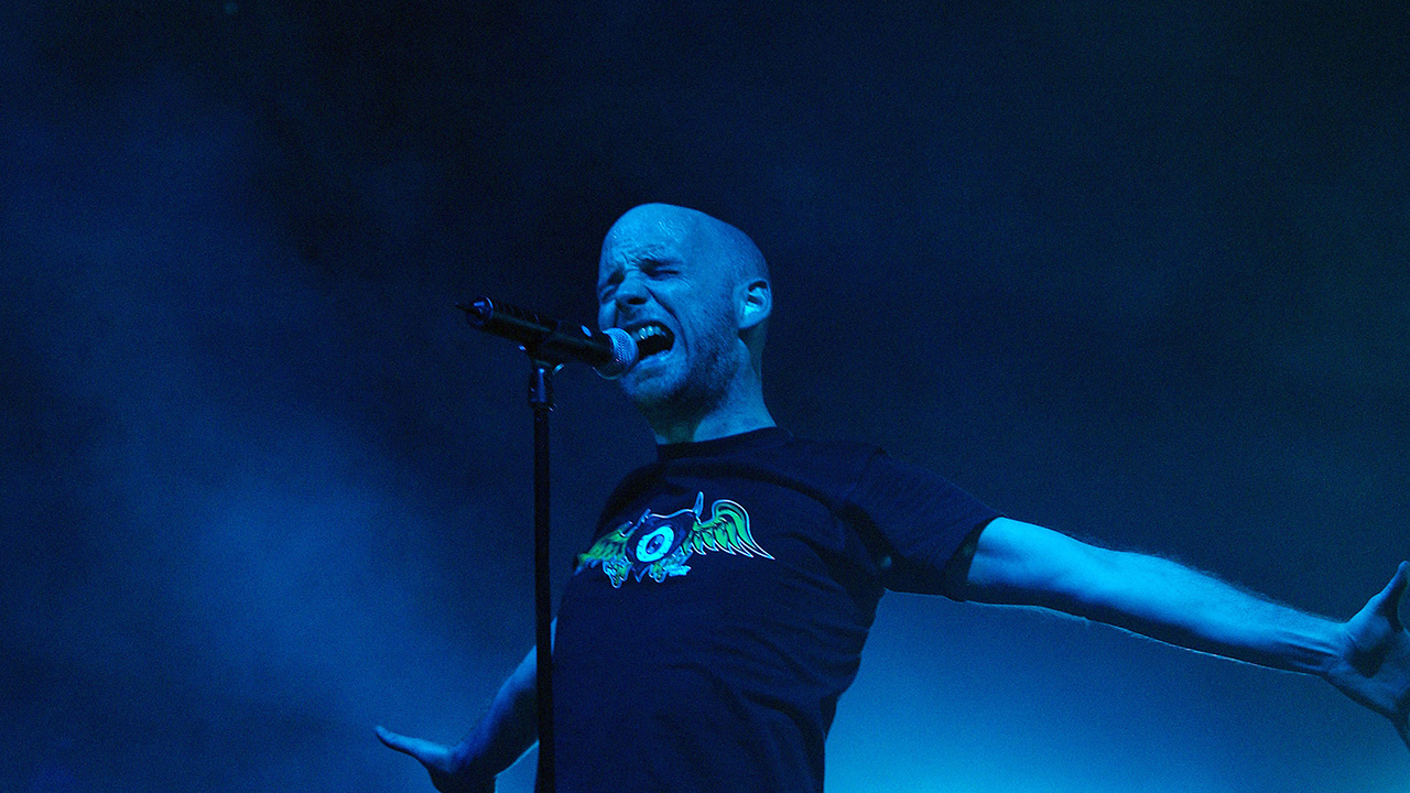 Moby performs on stage at a concert in 2002. He is in front of a mic in a black t-shirt with a weird eyeball design on it. He's sort of pushing forward with his torso as his arms go backward.