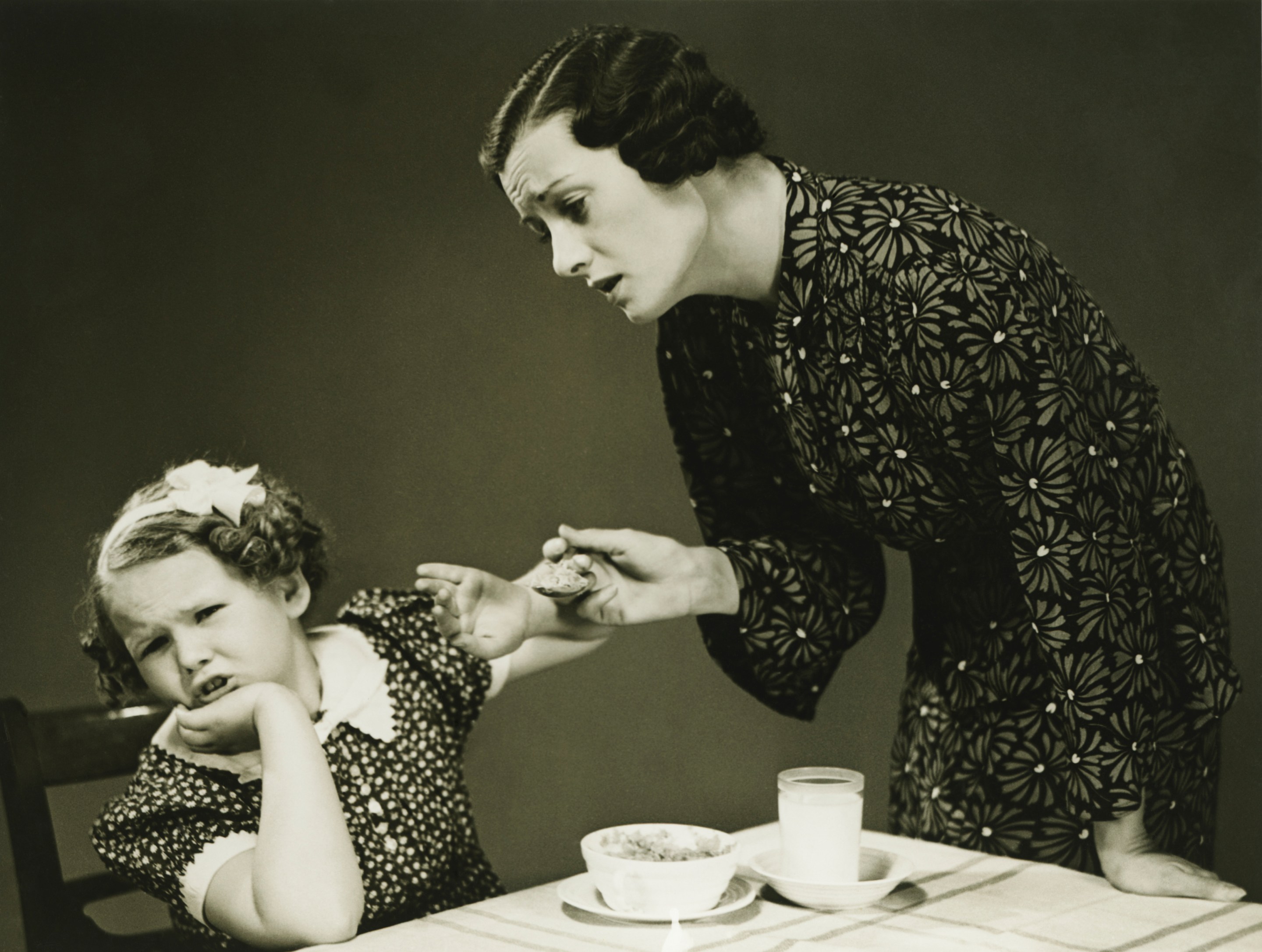 Mother trying to make daughter (4-5) finish food at table. It's an old stock photo from the 1940s.