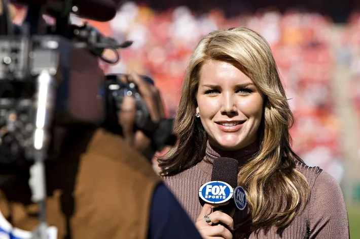 Charissa Thompson of Fox Sports seen talking into a microphone with a TV camera pointed at her.