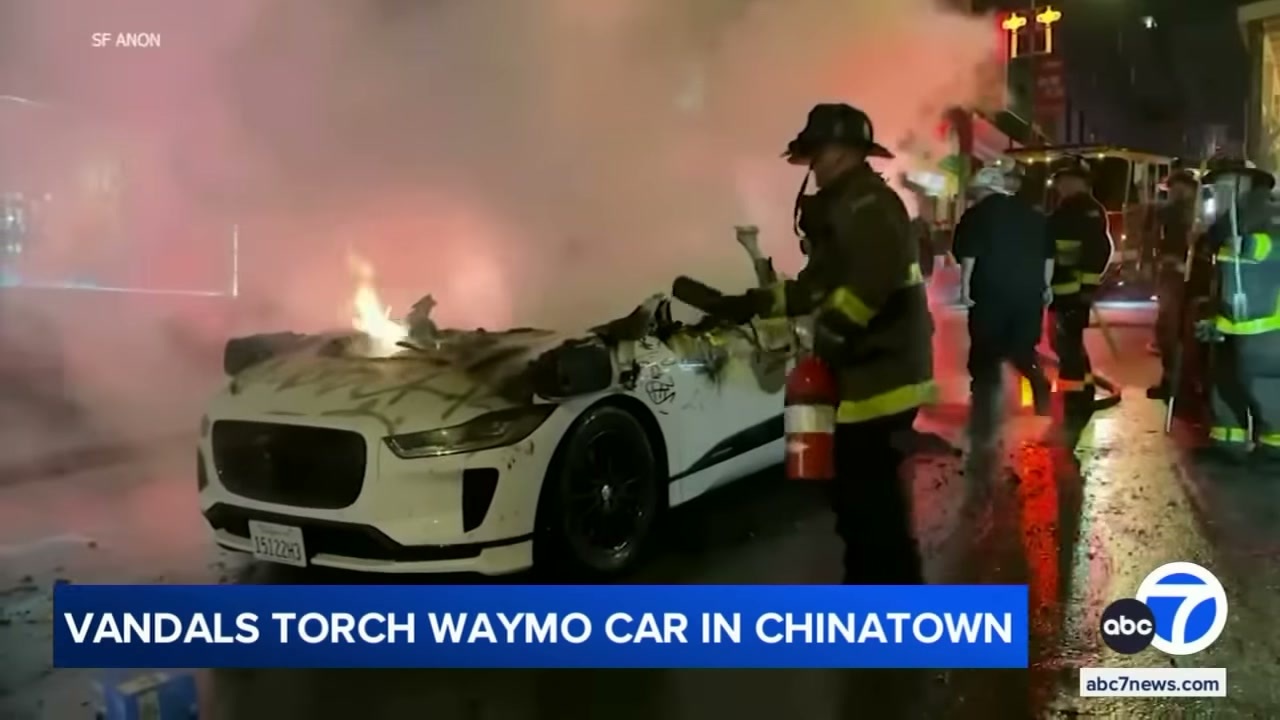 Firemen extinguish a Waymo autonomous vehicle that was set on fire by a crowd in San Francisco. It's a screengrab of news footage.