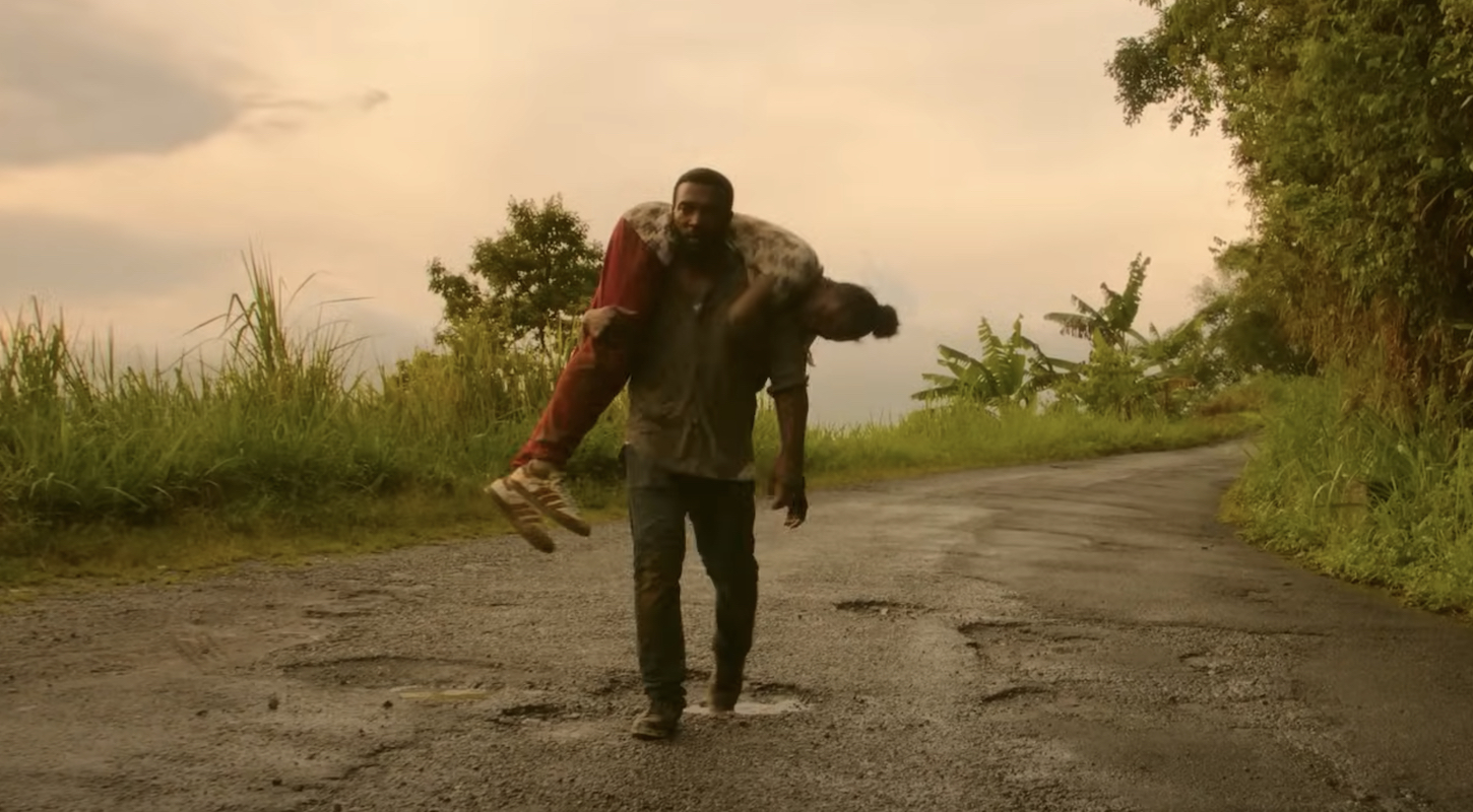 A man walks on a pockmarked road, carrying another man across his shoulders, from the TV series 'Top Boy.'
