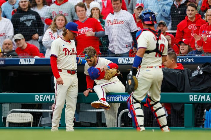 PHILADELPHIA, PENNSYLVANIA - MARCH 30: First baseman Bryce Harper #3 of the Philadelphia Phillies is helped out of the dugout by Bryson Stott #5 and J.T. Realmuto #10 after he chased a foul ball hit by Austin Riley #27 of the Atlanta Braves and flipped over the dugout railing during the first inning of a game at Citizens Bank Park on March 30, 2024 in Philadelphia, Pennsylvania.