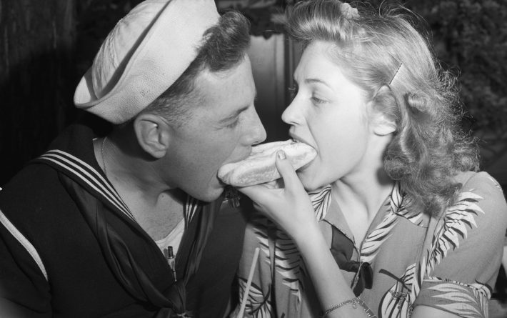 A woman and her sailor boyfriend eat from both ends of a hot dog. Coney Island.