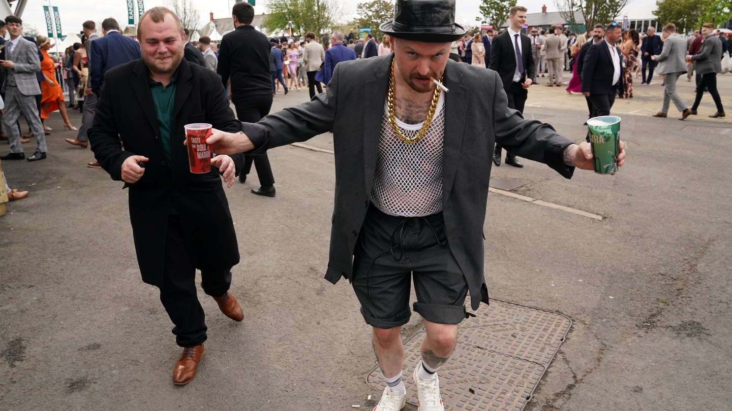 Racegoers on day two of the 2024 Randox Grand National Festival at Aintree Racecourse, Liverpool. One has cigarette in mouth, a drink in each hand, a heavy gold chain, and a mesh shirt.