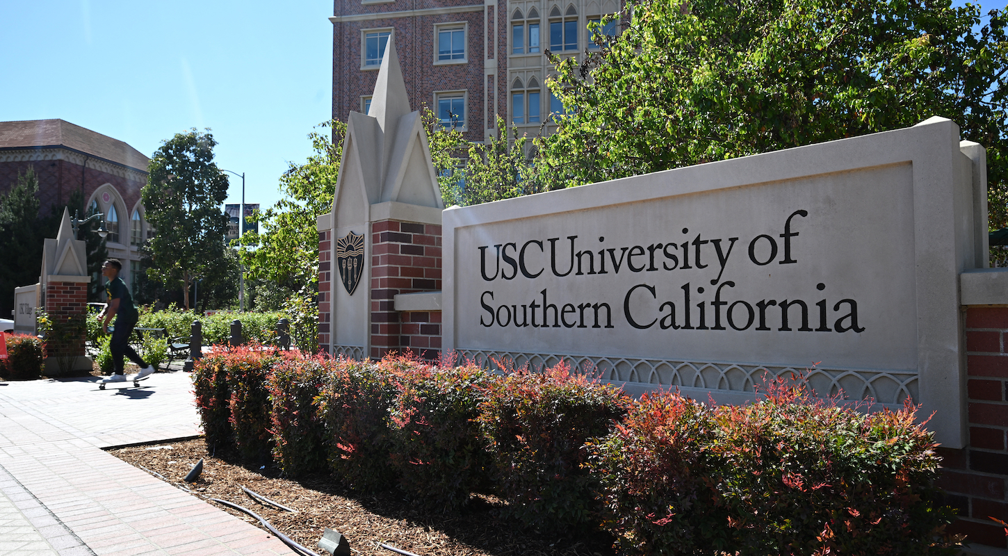 A big outdoor sign reading USC University of Southern California, with a student riding a skateboard in the background.
