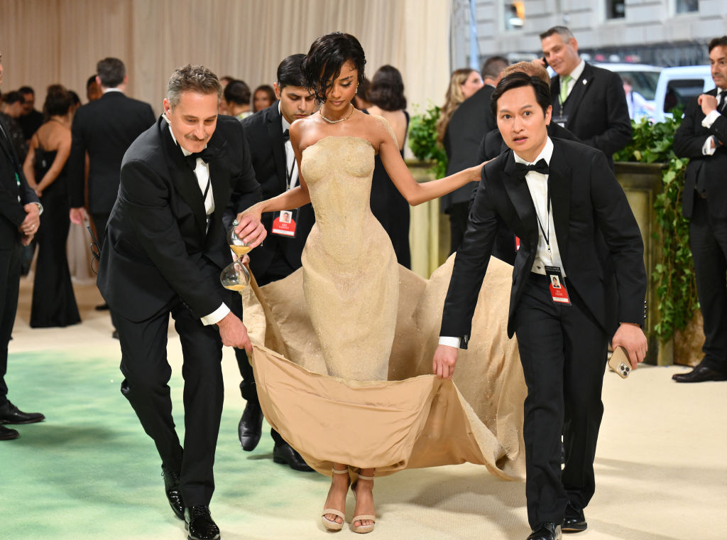 South African singer and songwriter Tyla arrives for the 2024 Met Gala in a Balmain gown meant to evoke sand in an hourglass. She is being helped by a procession of men in tuxedos who are holding the train of her gown in order for her to walk.