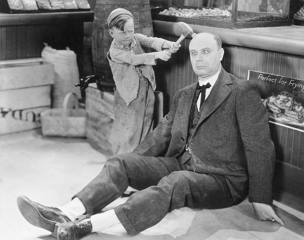 MOVIE GAG: CHILD ACTOR BOBBY 'WHEEZER' HUTCHINS OF THE OUR GANG TROUPE HITTING A MAN OVER THE HEAD WITH A HAMMER.UNDATED PHOTOGRAPH, CIRCA 1920s.