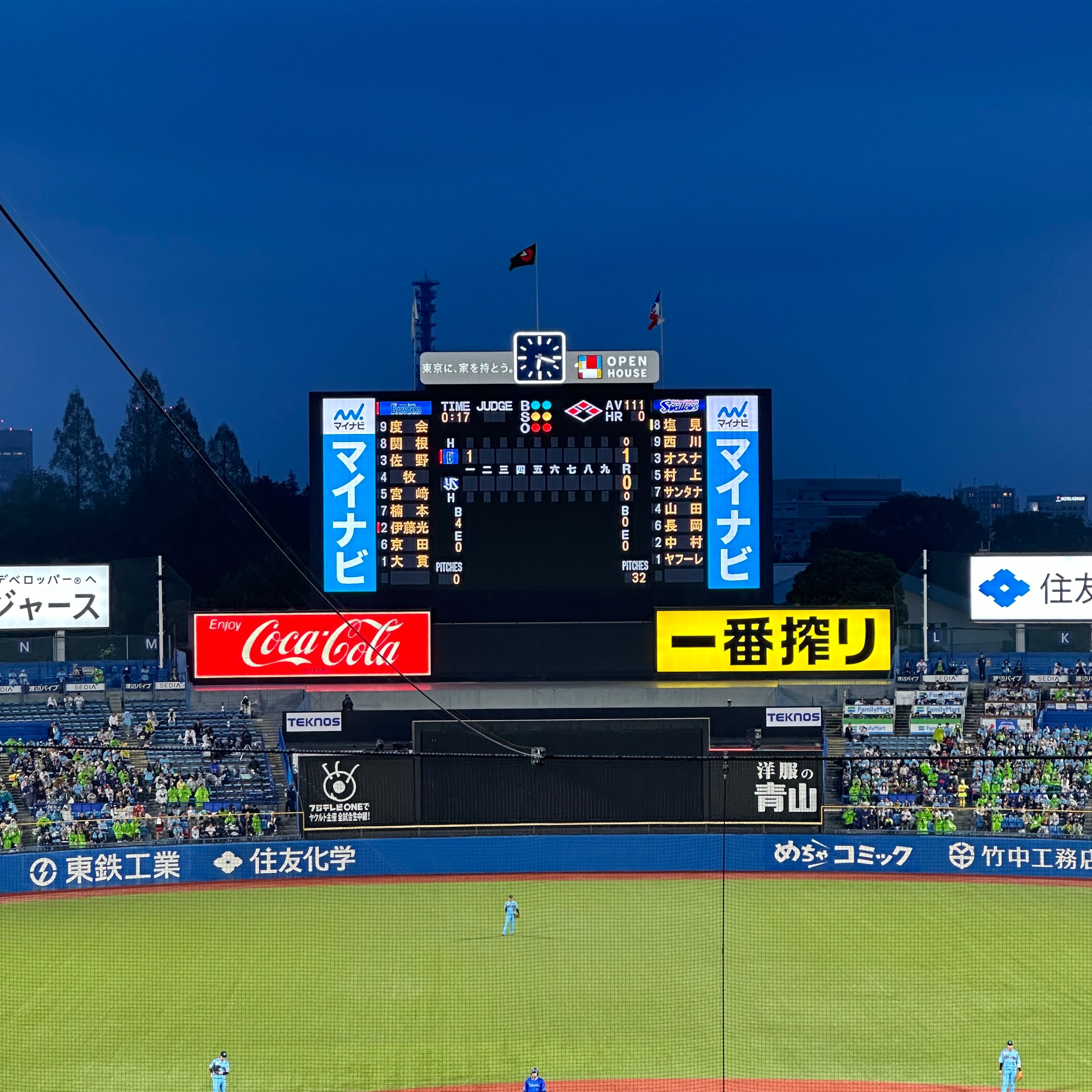 A photo of the baseball field and the scoreboard at Meji Jingu Baseball Stadium stadium in Tokyo. The night sky is a dark blue and, despite the rain, the outfield seats still hold a good number of fans. There's also a runner on second base.