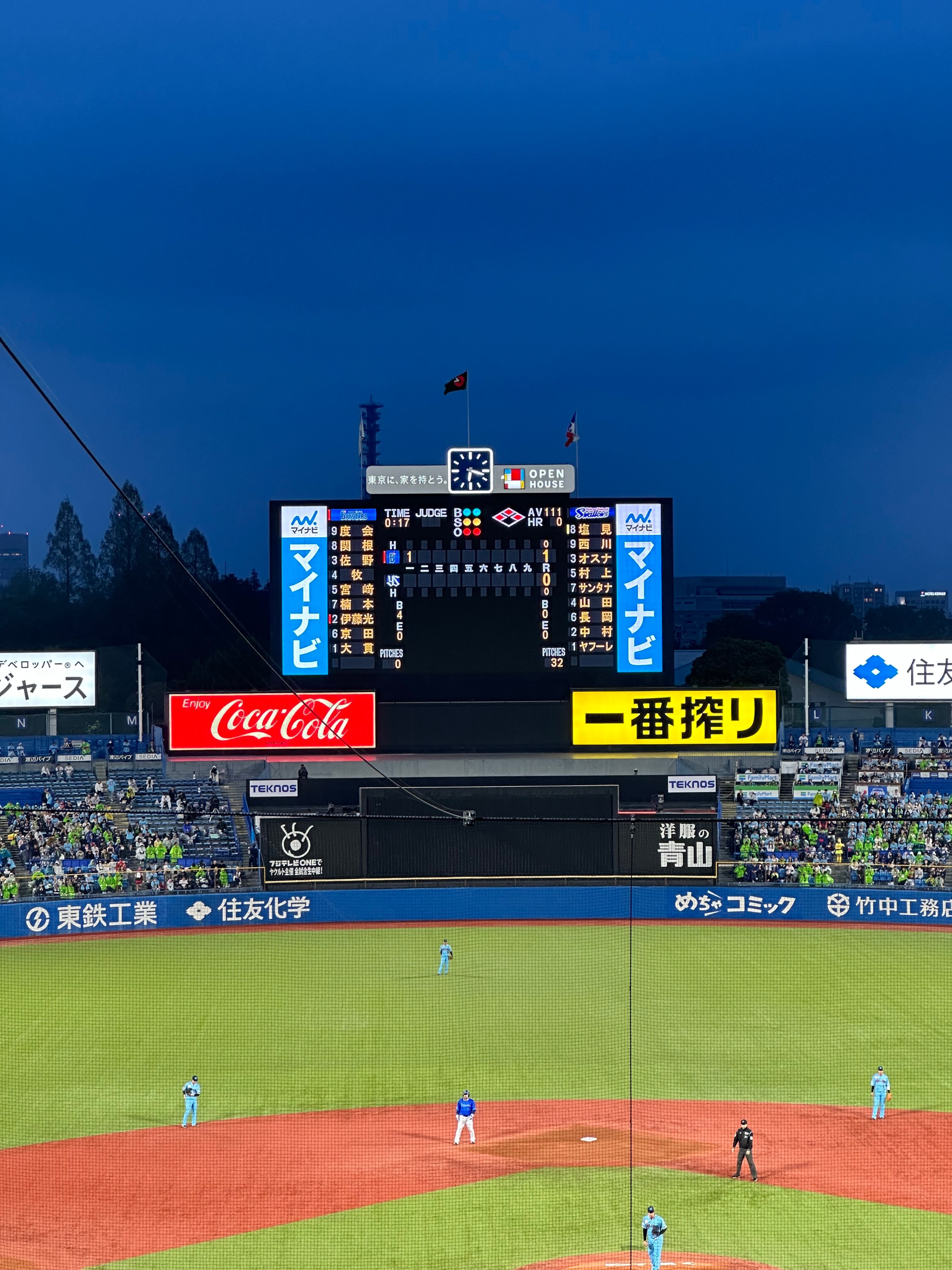 A photo of the baseball field and the scoreboard at Meji Jingu Baseball Stadium stadium in Tokyo. The night sky is a dark blue and, despite the rain, the outfield seats still hold a good number of fans. There's also a runner on second base.