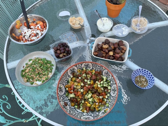 Various foods in serving dishes on a glass tabletop: Snap pea salad, shiraz salad, carrot and citrus salad, roasted potatoes, olives, and spreads.
