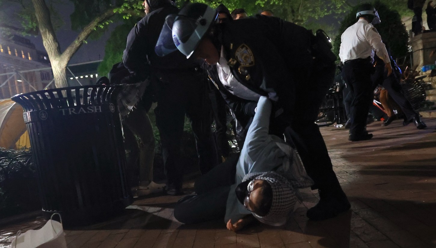 An NYPD officer in riot gear looms over a student protestor on the ground and appears to be gripping her arm.