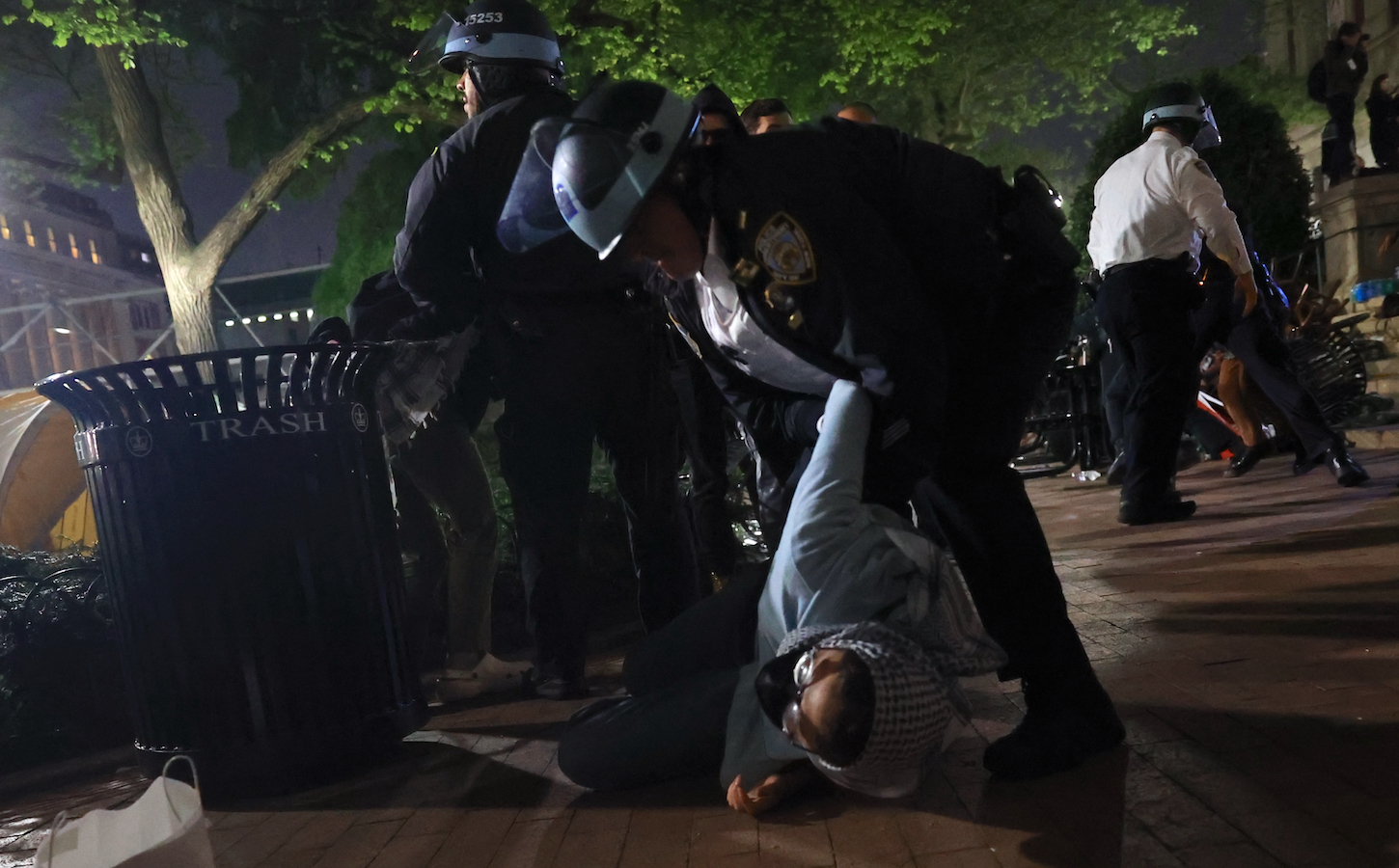 An NYPD officer in riot gear looms over a student protestor on the ground and appears to be gripping her arm.