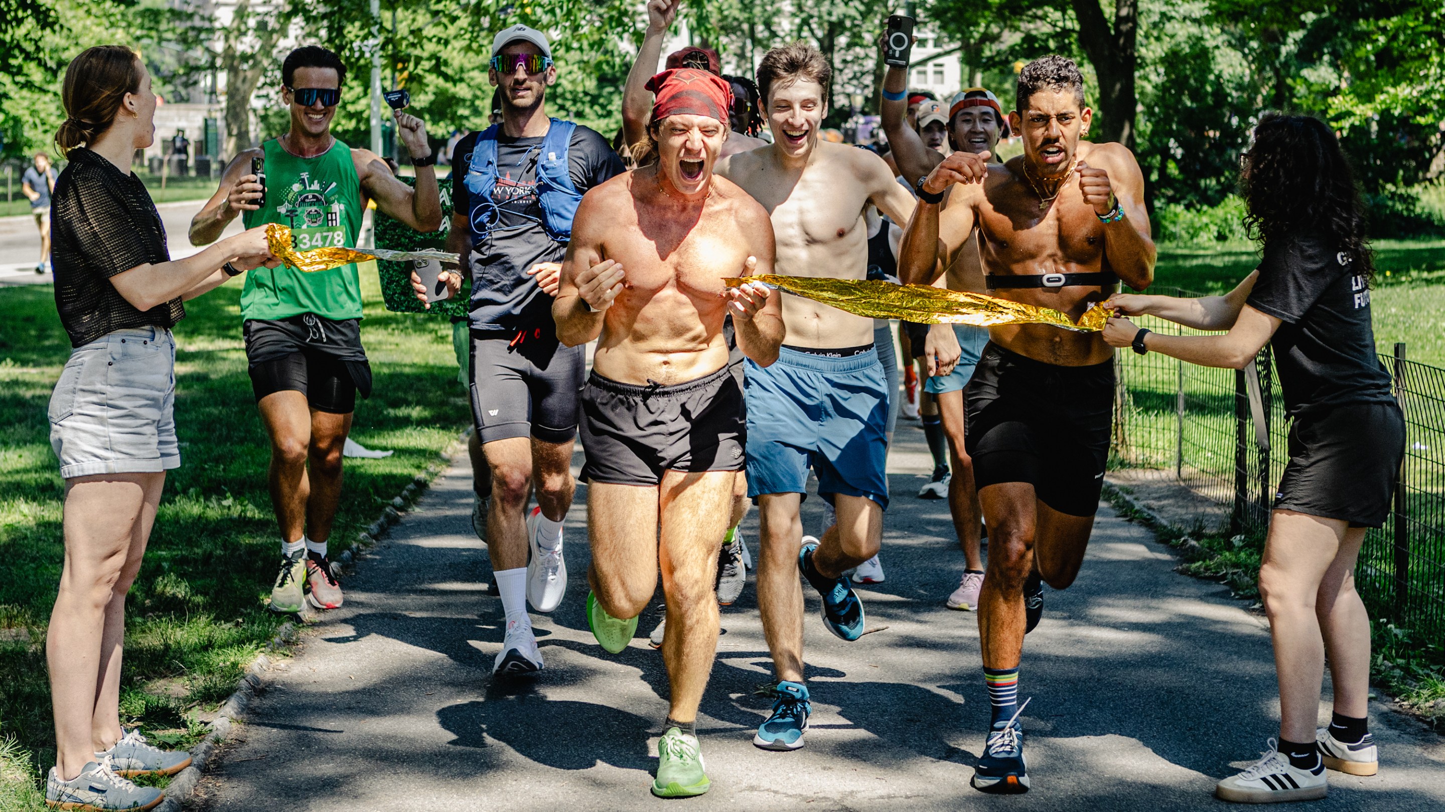 Kenny Moll rips through an impromptu finish line to mark the end of his seventh marathon. Michael-Luca is running to his right, with a crowd of runners behind them. There are trees in the background, and the area is both shaded and sunny.