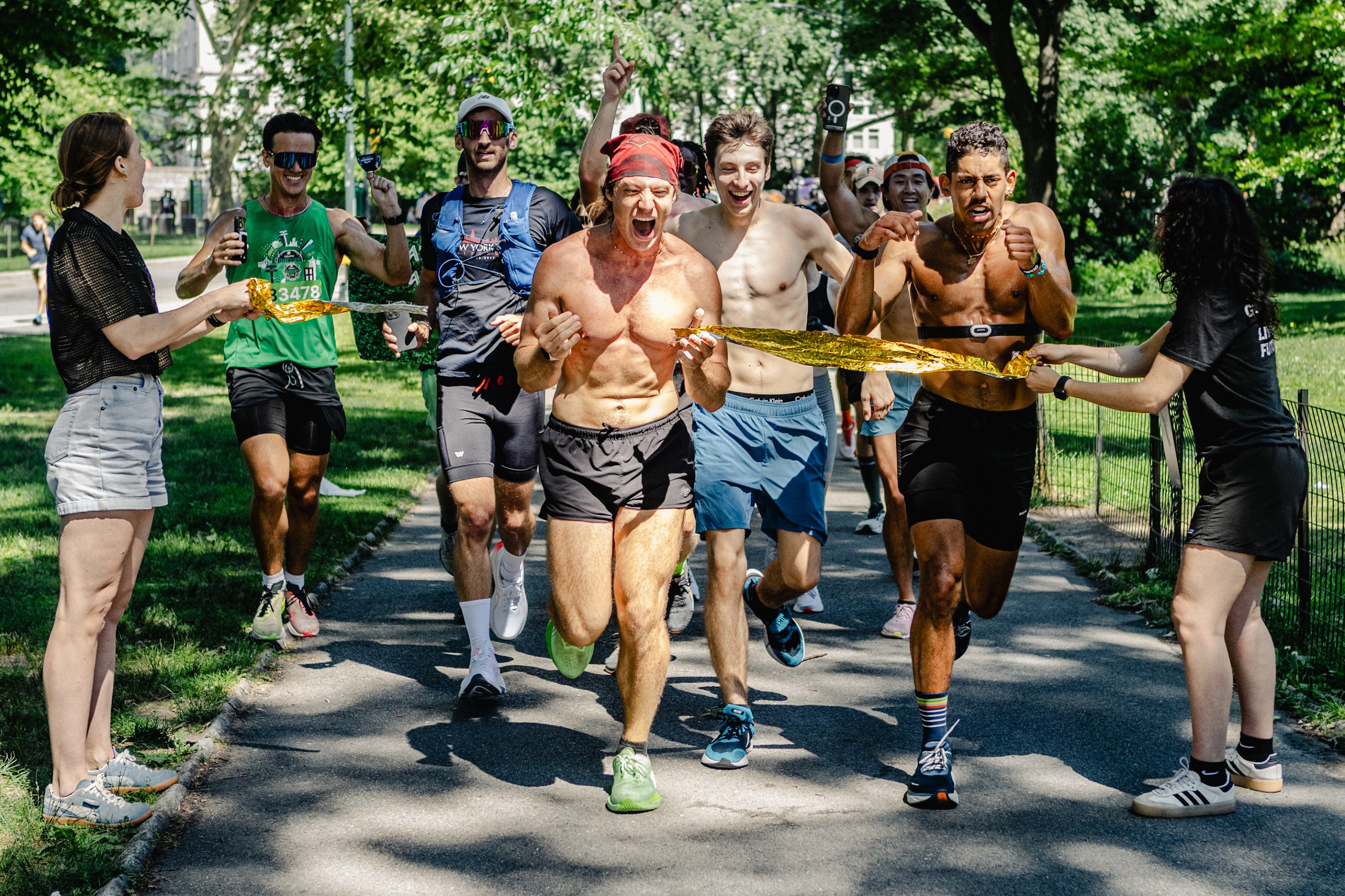 Kenny Moll rips through an impromptu finish line to mark the end of his seventh marathon. Michael-Luca is running to his right, with a crowd of runners behind them. There are trees in the background, and the area is both shaded and sunny.