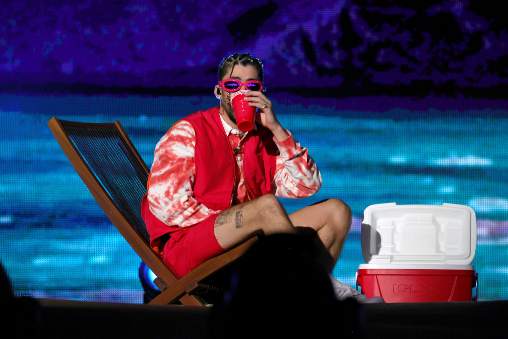 Bad Bunny performing on stage in Philadelphia in 2022, wearing an outfit with red shorts while sitting on a beach chair while enjoying a beverage.
