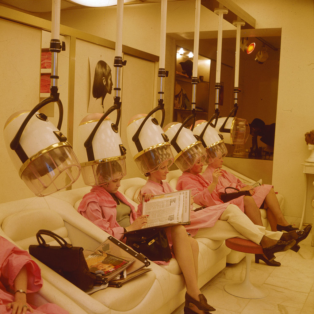 Women sit under hair dryers in a Paris beauty shop, smoking and reading magazines while their hair dries.