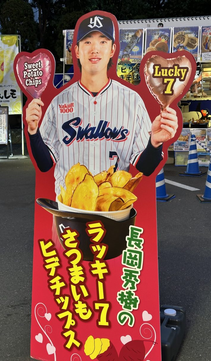 A giant cutout a Tokyo Yakult Swallows player in an advertisement for sweet potato chips. in one hand, he is holding a heart-shaped balloon that says "Sweet Potato Chips." The other hand, he is holding a heart-shaped balloon that says, "Lucky 7."