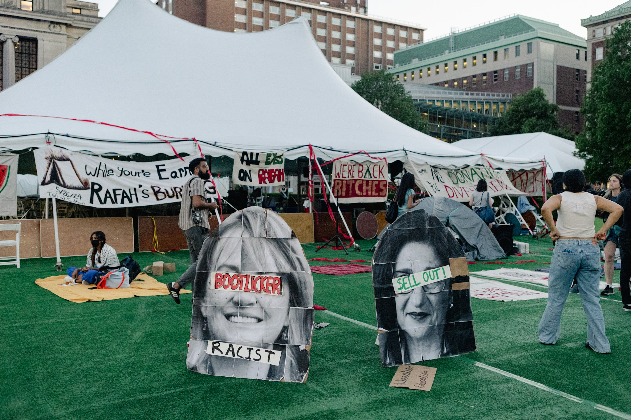 A photo of the west side of the South Lawn in front of Columbia University's Butler Library at the Morningside campus. This picture, taken right before sunset, depicts an encampment set up by protestors on a lawn with turf and a big event tent. Students are scattered throughout, and there are multiple banners and signs depicting pro-Palestine statements.