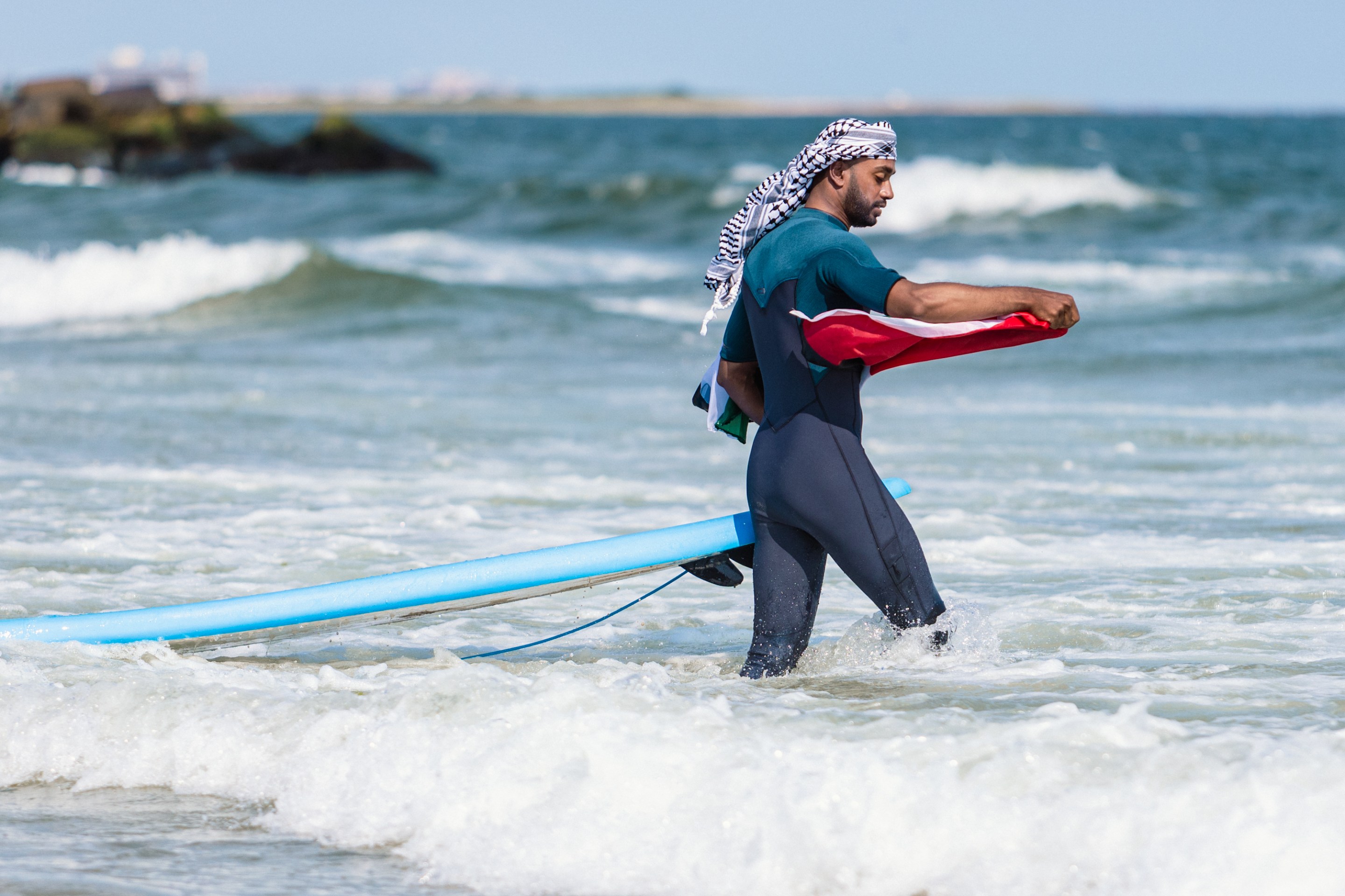 A surfer in a blue wetsuit carries a light blue surfboard with him as he walks into the ocean. He is holding a Palestinian flag in his right hand and wearing a keffiyeh around his head.