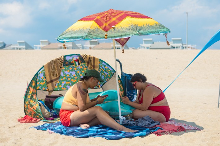 Two beachgoers sit on a beach towel on a sandy beach. They have an umbrella over them and a beach shade tent behind them. They are both on their phones.