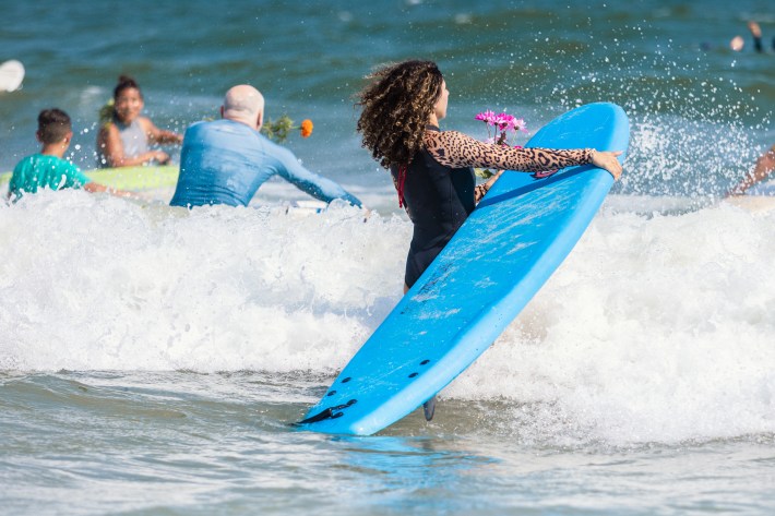 A surfer with a blue surf board walks into the ocean while holding pink flowers for the paddle out.