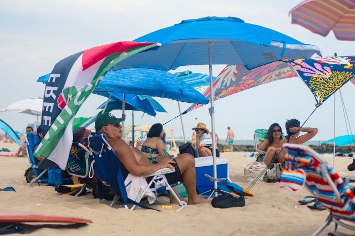 A man lounges in a beach chair on the beach under a blue umbrella with a "Free Palestine" flag attached to it. There is a blue cooler next to him and other beachgoers around him sitting in beach chairs.